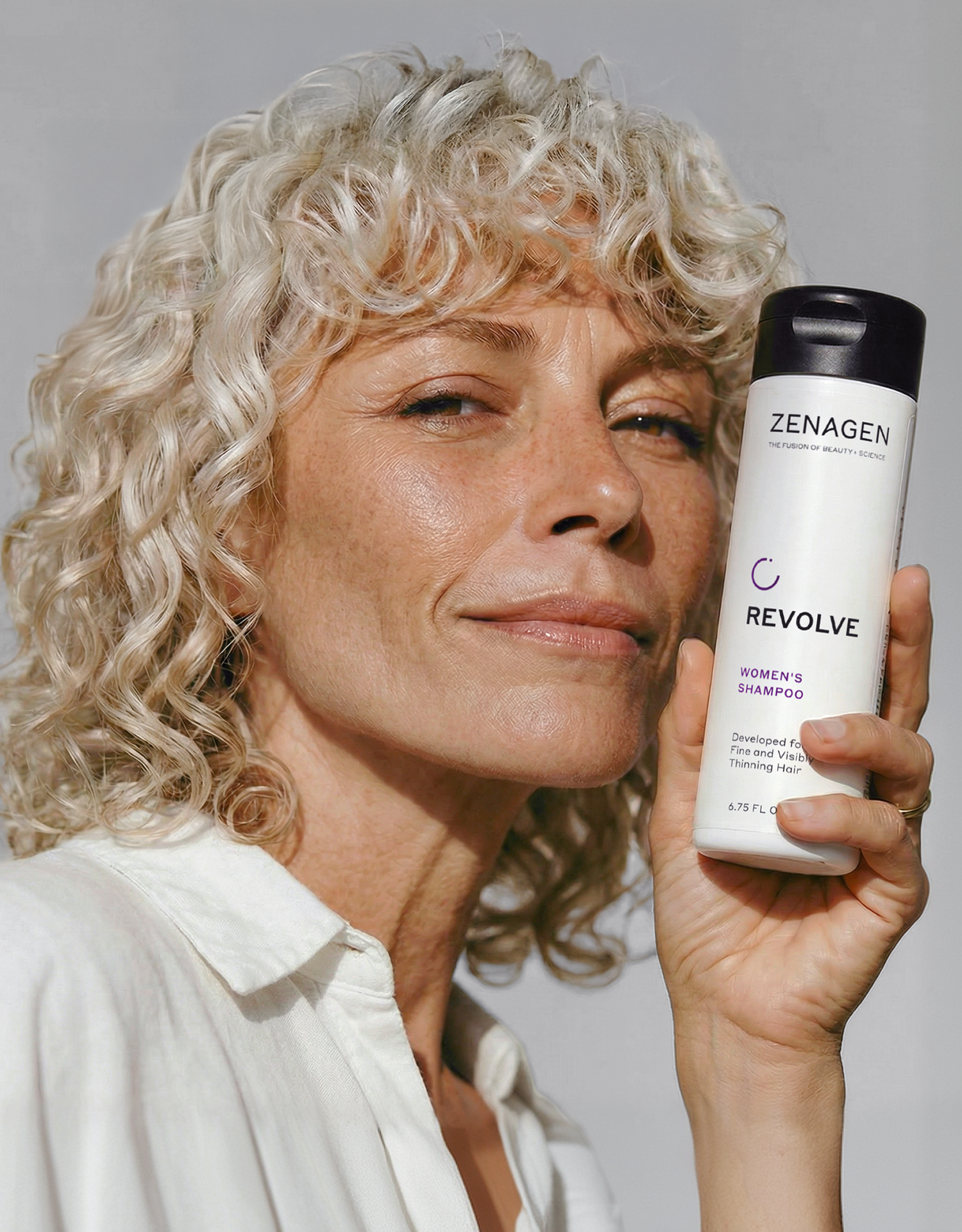 Woman with curly blonde hair holding Zenagen Revolve women’s shampoo for fine and thinning hair, supporting a healthy-looking scalp and fuller appearance
